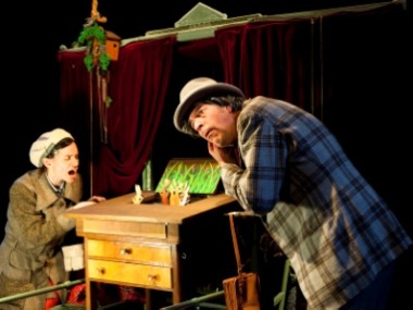 © Theater Mummpitz Michael Bang horcht auf einer Theater-Bühne vor einem roten Samt-Vorhang nach Tönen, die aus einem aufklappbaren Tisch zu kommen scheinen