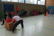 Zwei Kinder sitzen in einem umgedrehten Kasten, der auf einem Rollbrett steht, und werden von zwei weiteren Kindern geschoben.