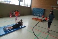 Kinder aktiv in der Sporthalle Ein Kind liegt in einer Sporthalle auf der Matte und vier bewegeb sich drumherum.