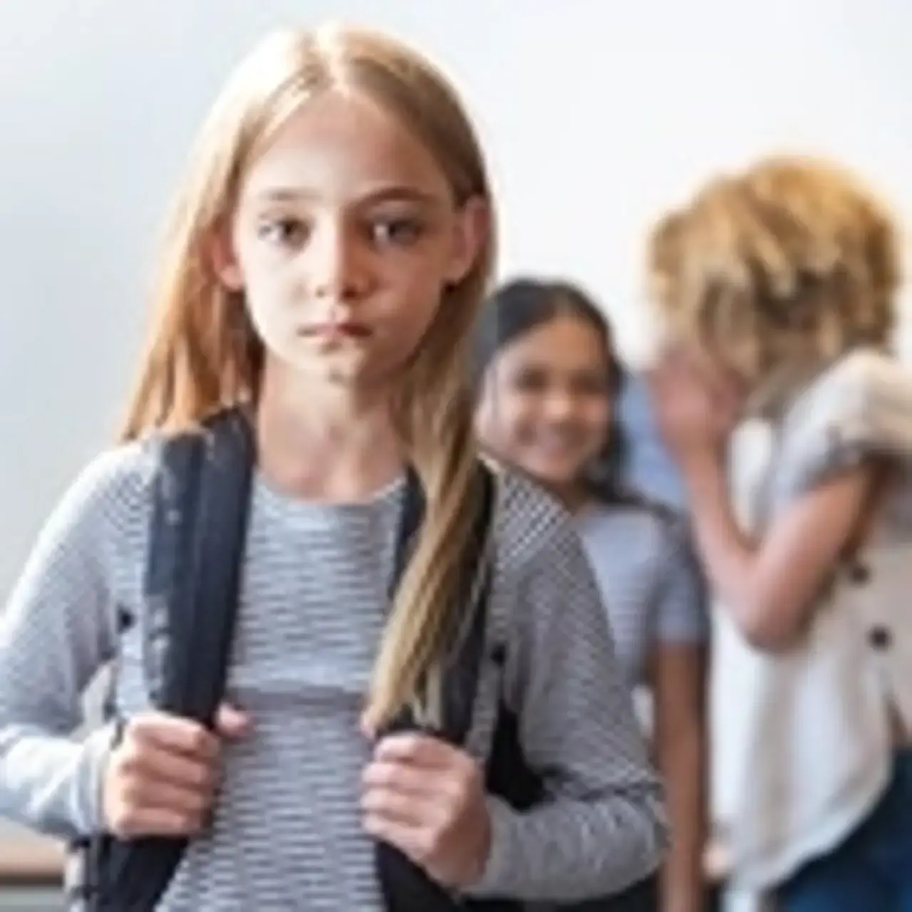 Traurig aussehendes Mädchen im Vordergrund. Im Hintergrund: Kinder, die hinter vorgehaltener Hand tuscheln.