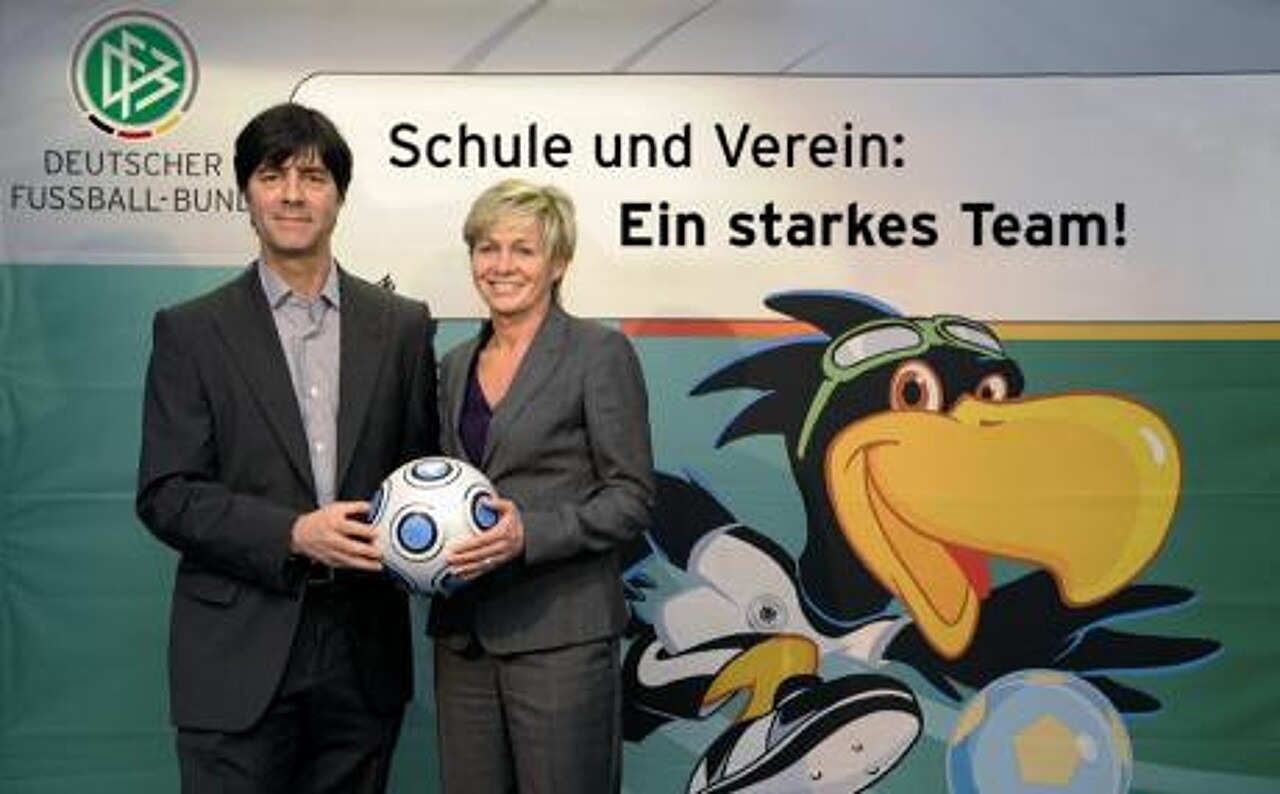Die Bundestrainer Joachim Löw und Silvia Neid stehen gemeinsam vor einer DFB-Fotowand mit dem Schriftzug „Schule und Verein: Ein starkes Team!“ und halten dabei gemeinsam einen Fußball