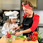 Frau Güßbacher von kinder-kochkurs.com und ein Mädchen mit Kochmütze schnibbeln in der Küche Gemüse.