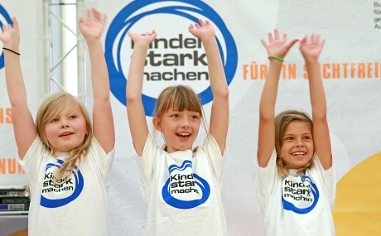 Drei Mädchen strecken lächeln die Arme vor einer „Kinder stark machen“-Fotowand in die Luft