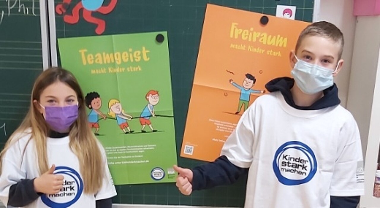 Zwei Kinder mit einem Kinder stark machen T-Shirt stehen vor den Plakaten 