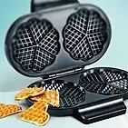  Doppel-Waffeleisen in klassischer Herzform mit frisch gebackenen Waffeln daneben.