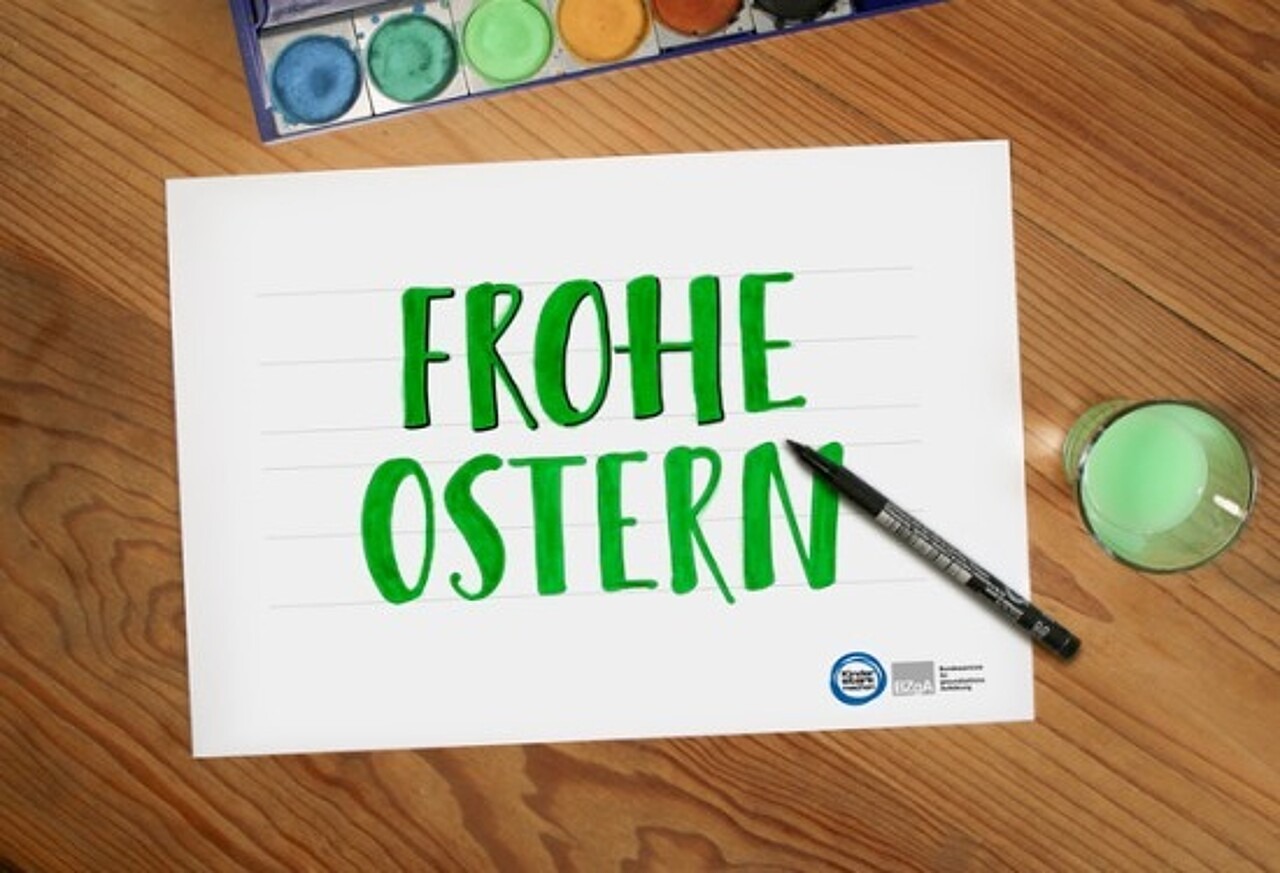 ausgemalte Vorlage mit Schriftzug „Frohe Ostern“, daneben Farbkasten, Glas mit grün gefärbtem Wasser und ein schwarzer Fineliner