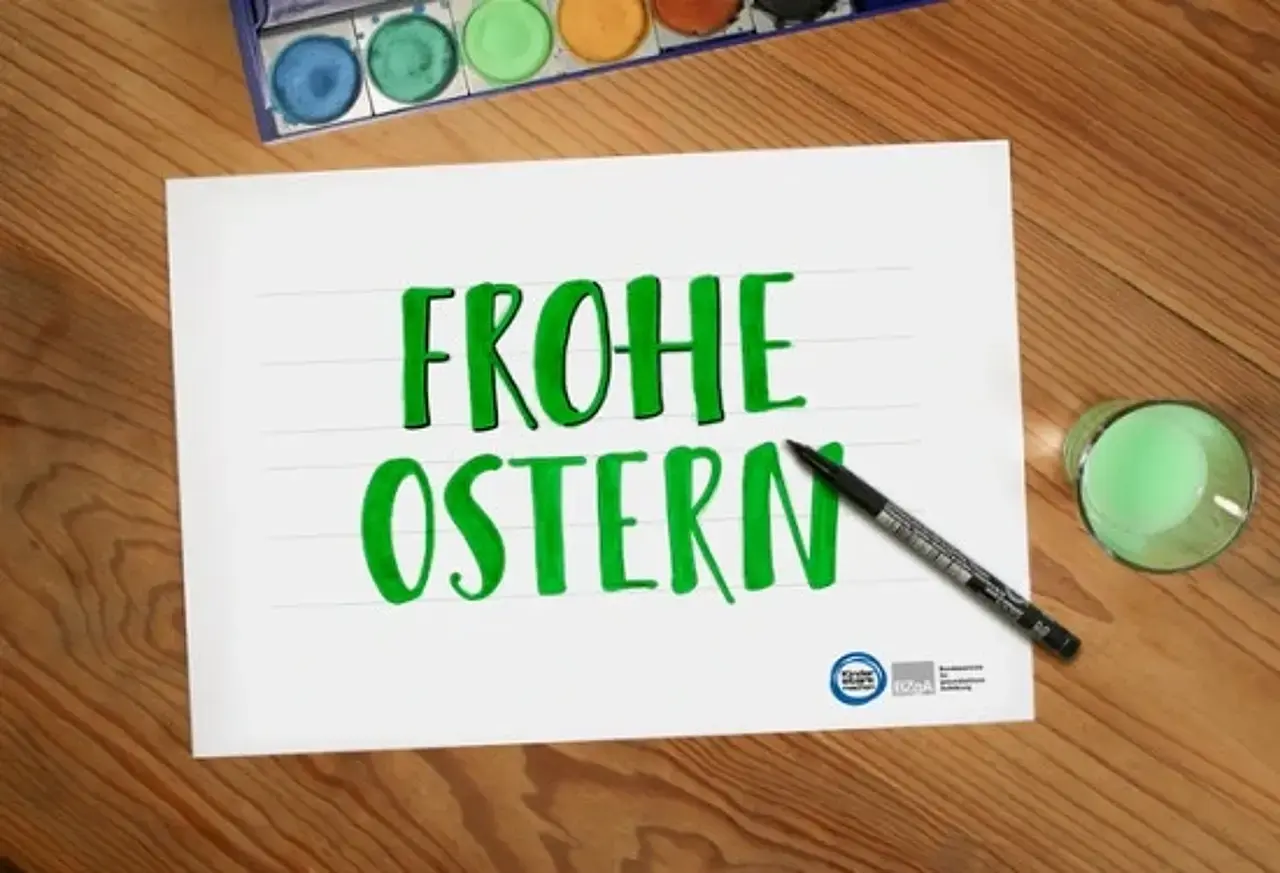 ausgemalte Vorlage mit Schriftzug „Frohe Ostern“, daneben Farbkasten, Glas mit grün gefärbtem Wasser und ein schwarzer Fineliner
