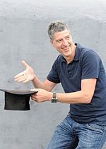 Michael Bang in Jeans und dunkelblauem Polo-Shirt zaubert mit einem Zylinder-Hut vor einer grauen Wand
