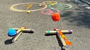 Zwei selbstgebaute Katapulte aus Holzstäbchen und Haushaltsgummis: Von einem fliegt ein Schwämmchen durch die Luft Richtung Kreide-Kreise auf der Straße.