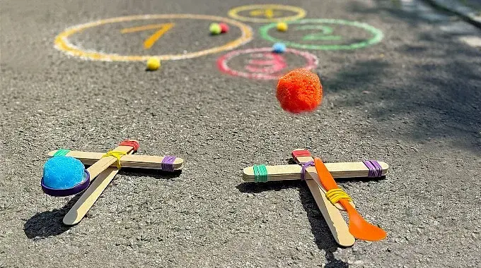 Zwei selbstgebaute Katapulte aus Holzstäbchen und Haushaltsgummis: Von einem fliegt ein Schwämmchen durch die Luft Richtung Kreide-Kreise auf der Straße.