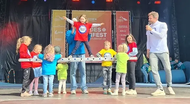 Kinder auf der Hauptbühne mit dem „Kinder stark machen“-Team. Die Kinder halten eine Leiter waagerecht und ein Mädchen steht freihändig auf der Leiter.