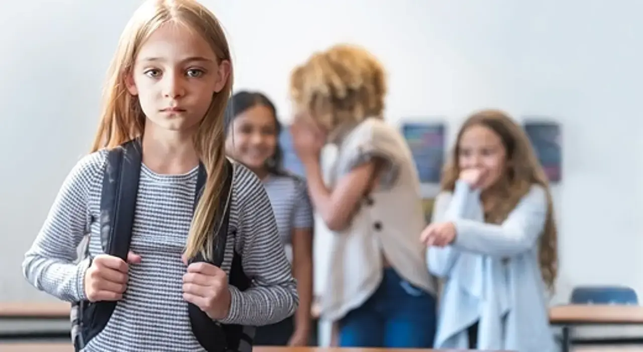 Traurig aussehendes Mädchen im Vordergrund. Im Hintergrund: Kinder, die hinter vorgehaltener Hand tuscheln.