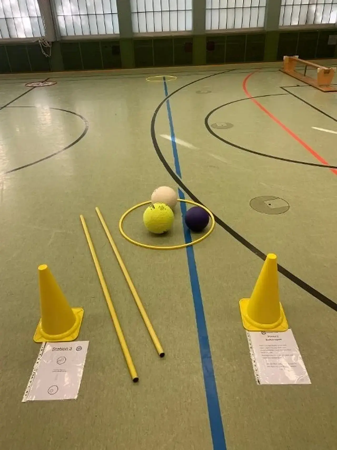 2 Reifen mit 10m-Abstand auf dem Hallenbogen, in 1 Reifen 3 Bälle und daneben zwei Stäbe. daneben zwei Stäbe