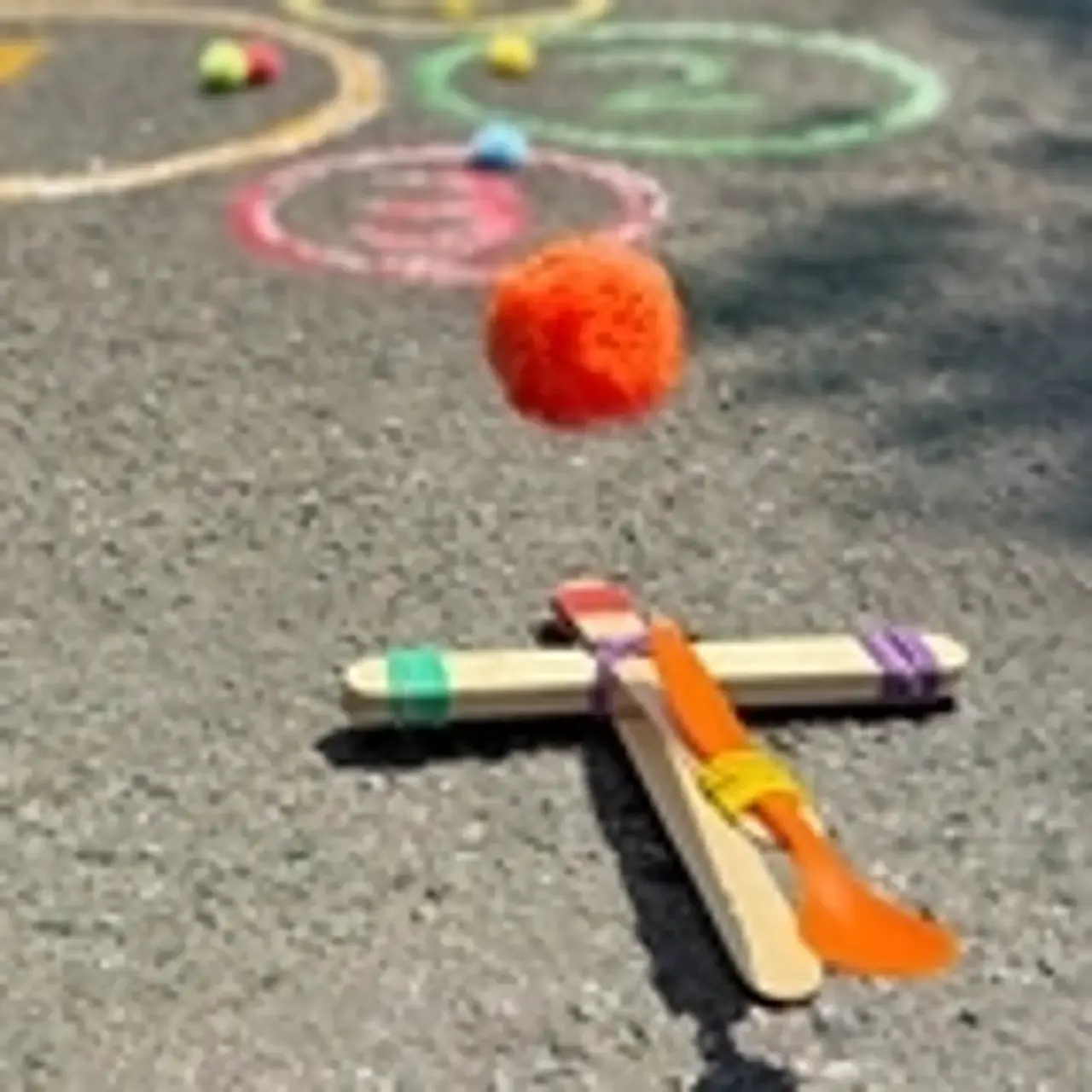 Ein selbstgebautes Katapult aus Holzstäbchen und Haushaltsgummis: Von einem fliegt ein Schwämmchen durch die Luft Richtung Kreide-Kreise auf der Straße.