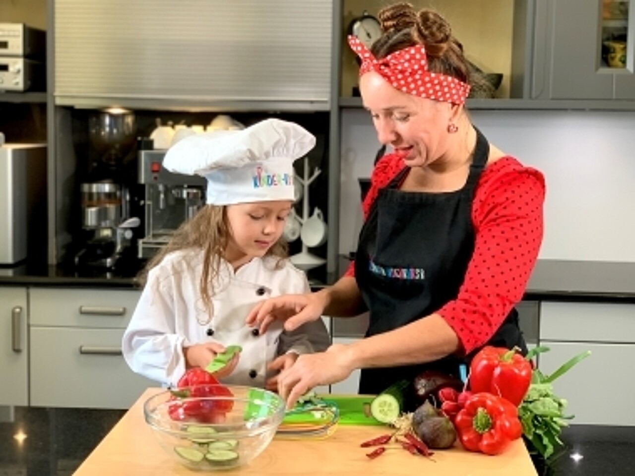Frau Gueßbacher von kinder-kochkurs.com und ein Mädchen mit Kochmütze schnibbeln in der Küche Gemüse.