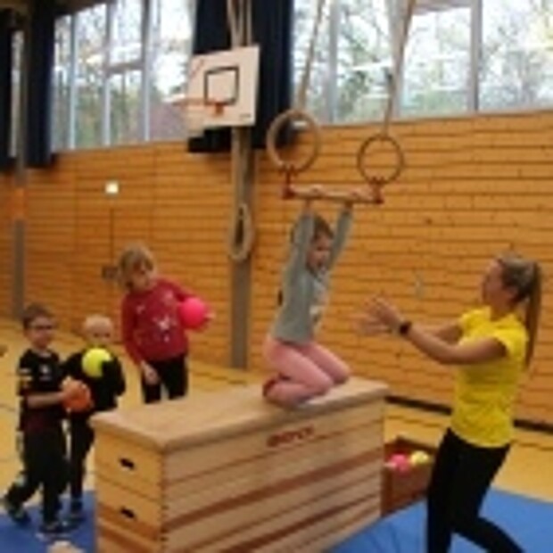 Kinder an Bewegungsstation: Ein Mädchen schwingt mittels einer Ringstange über einen Kasten. Sie erhält dabei von ihrer Trainerin Hilfestellung.
