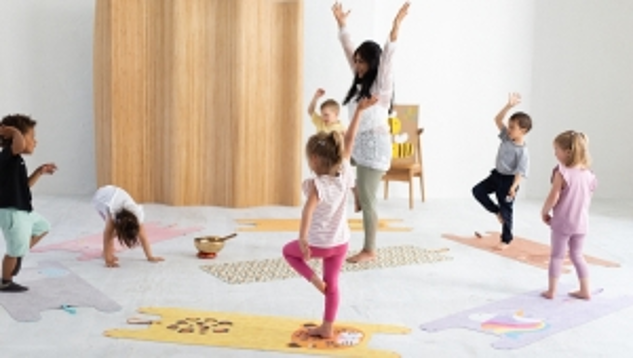 Kinder stehen im Kreis mit ihrer Betreuerin auf Yogamatten mit Tiermotiven und üben sich im Einbeinstand.