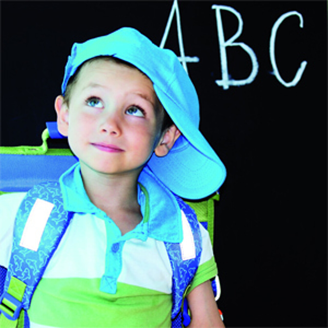 kleiner Junge mit Ranzen und Kapuzenmütze steht vor einer schwarzen Tafel, auf der ABC geschrieben steht