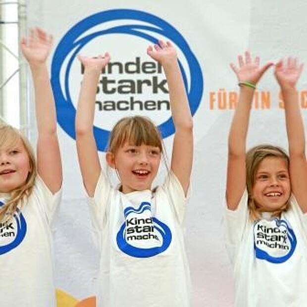 Drei Mädchen strecken lächelnd ihre Arme vor einer „Kinder stark machen“-Fotowand in die Luft