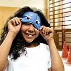 Ein Mädchen in der Turnhalle hebt die Augenbinde leicht an und lächelt darunter hervor.