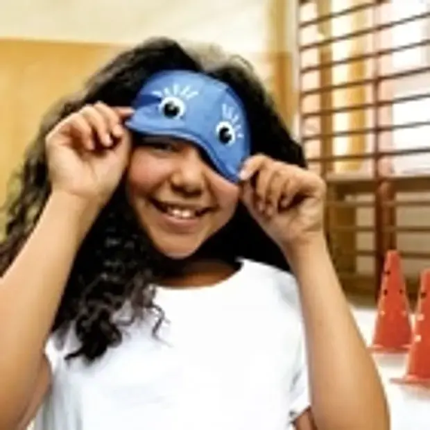 Ein Mädchen in der Turnhalle hebt die Augenbinde leicht an und lächelt darunter hervor.