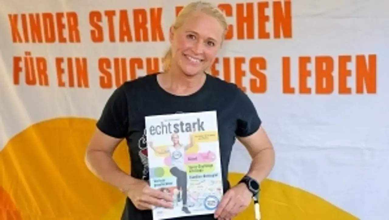 Singa Gätgens mit dem Mitmachheft in der Hand. Im Hintergrund dre Slogan: "Kinder stark machen für ein suchtfreies Leben"