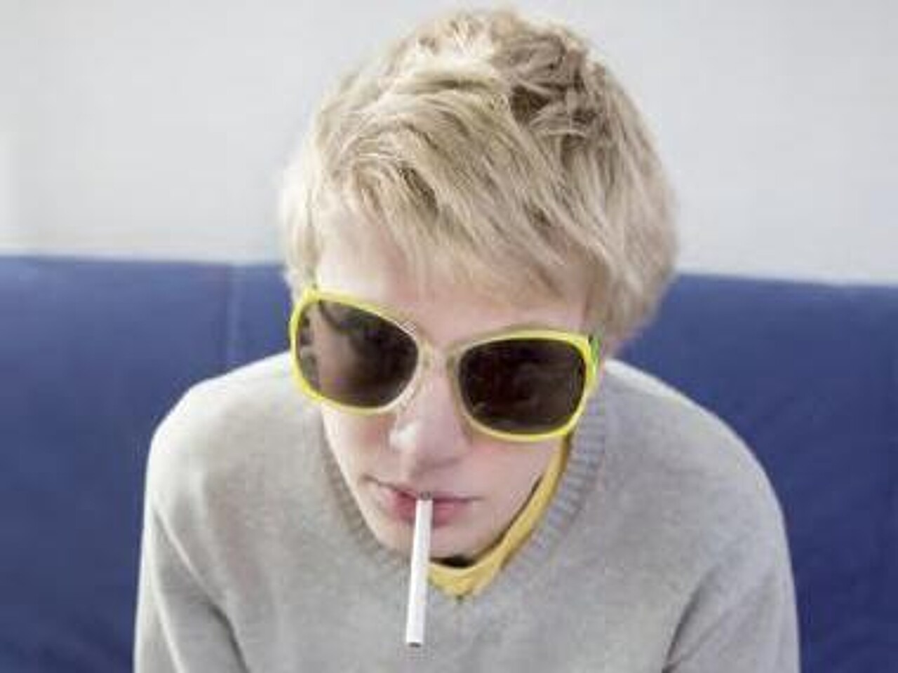 Junge mit gelb umrandeter Sonnenbrille sitzt auf einem Sofa und hat eine Zigarette im Mund