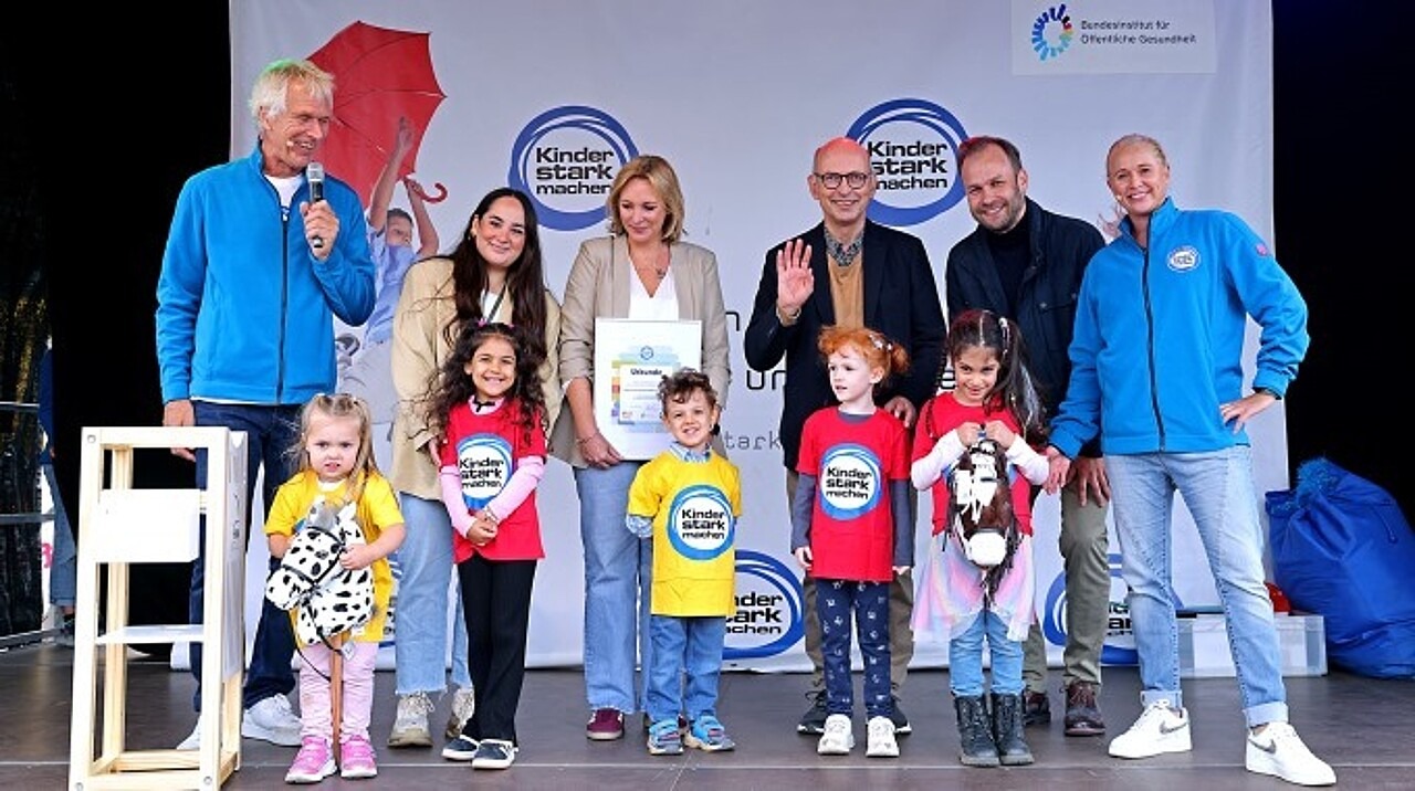 Dezernent Robert Voigtsberger und der kommissarische BIÖG-Leiter Dr. Johannes Nießen übergeben den Anerkennungspreis an die Leiterin der Kita Hunoldstraße im Beisein vieler Kinder