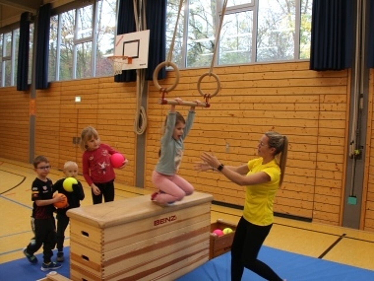 Kinder an Bewegungsstation: Ein Mädchen schwingt mittels einer Ringstange über einen Kasten. Sie erhält dabei von ihrer Trainerin Hilfestellung.