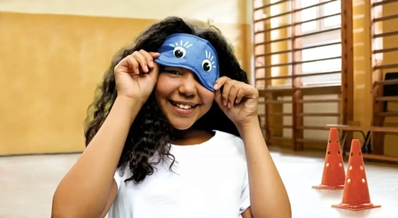 Ein Mädchen in der Turnhalle hebt die Augenbinde leicht an und lächelt darunter hervor.