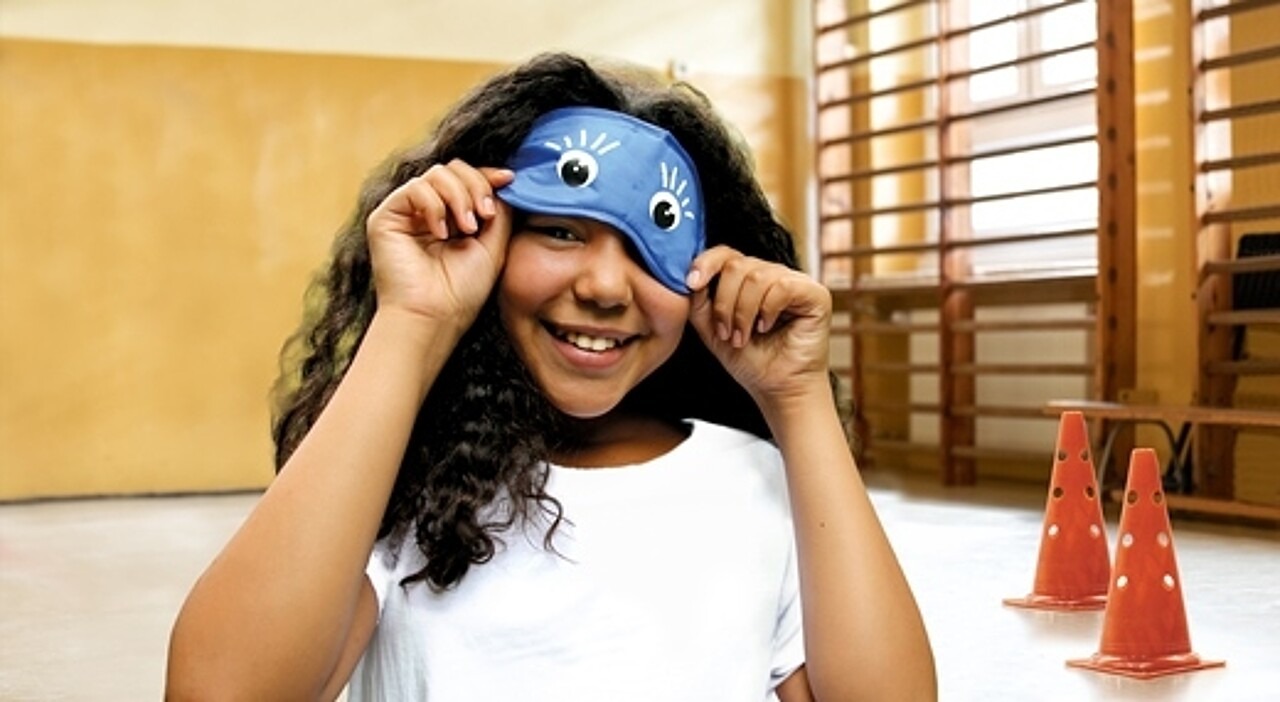 Ein Mädchen in der Turnhalle hebt die Augenbinde leicht an und lächelt darunter hervor.