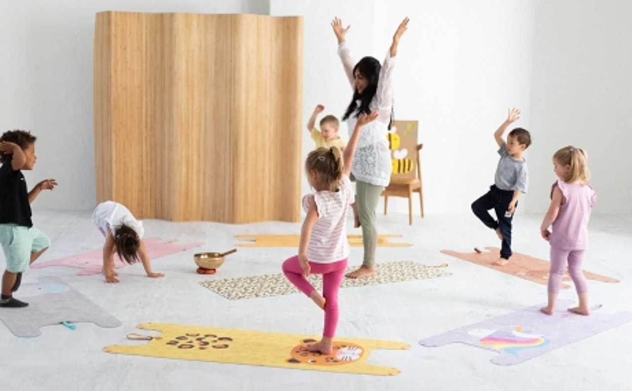 Kinder stehen im Kreis mit ihrer Betreuerin auf Yogamatten mit Tiermotiven und üben sich im Einbeinstand.