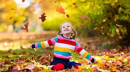 Ein Mädchen sitzt im leuchtenden Herbstlaub und wirft die Blätter in die Luft.