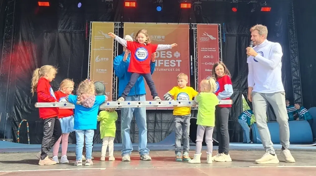Kinder auf der Hauptbühne mit dem „Kinder stark machen“-Team. Die Kinder halten eine Leiter waagerecht und ein Mädchen steht freihändig auf der Leiter.