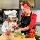 Frau Güßbacher von kinder-kochkurs.com und ein Mädchen mit Kochmütze schnibbeln in der Küche Gemüse.