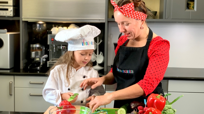 Frau Güßbacher von kinder-kochkurs.com und ein Mädchen mit Kochmütze schnibbeln in der Küche Gemüse.