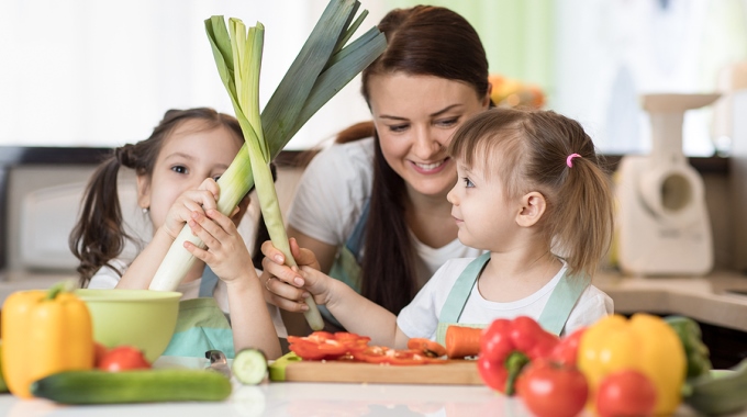 Eine Mutter mit ihren beiden kleinen Töchtern beim Schneiden von frischem Gemüse.